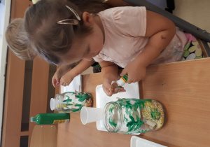 Dziewczynka przykleja rybki na pomalowany słoik.