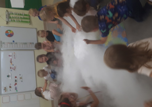 Dzieci siedzą wkole w chmurze powstałej przez dodanie suchego lodu do ciepłej wody.