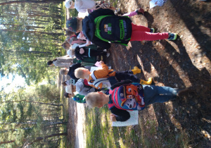 Grupa dzieci trzymając w ręku mapę idzie za przewodnikiem i szuka ukrytych w lesie informacji o jeżach.