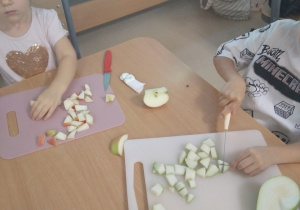 Dzieci siedzą przy stole I kroją owoce w kostkę na swojej desce do krojenia.