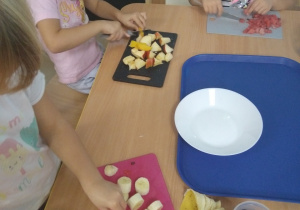 Dziewczynki siedzą przy stole i kroją owoce kostkę.