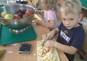 Dzieci siedzą przy stole I są skupione na krojeniu na swoich deskach różnych owoców.