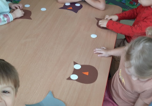 Kilkoro dzieci siedzi przy stoliku wykonując papierową sowę.
