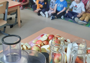 Dzieci siedzą na dywanie, na stoliku są owoce i sokowirówka