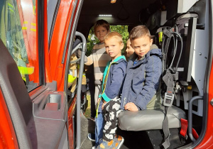 Trzech chłopców siedzi w wozie strażackim i pozuje do zdjęcia.