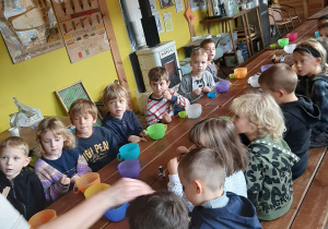 Dzieci siedzą przy stole, słuchają instrukcji wykonania mydełek glicerynowych