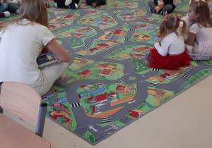Dzieci siedzą na dywanie trzymając pluszowe misie, słuchają bajki czytanej