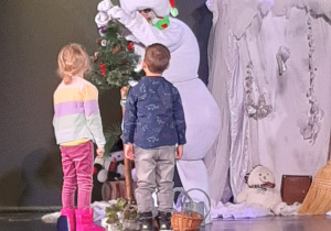 2 dzieci stoi na scenie, obok aktor przebrany za bałwana ubiera choinkę