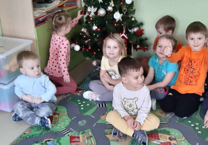 Dzieci siedzą na dywanie obok ubranej choinki