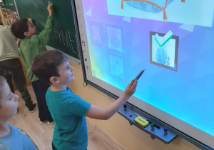 Chłopiec wykonuje zadanie na tablicy interaktywnej, a chłopiec na drugim planie pisze swoje imię na tablicy