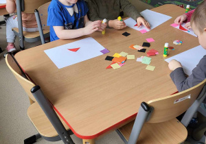 Dzieci siedzą przy stolikach i naklejają figury geometryczne na kartki