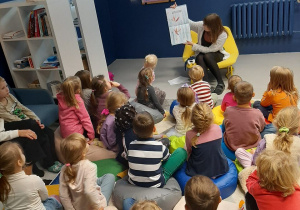 Dzieci siedzą na poduszkach i słuchają bajki czytanej przez Panią Bibliotekarkę