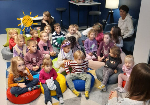 Dzieci siedzą na poduszkach i słuchają opowiadania czytanego przez Panią Bibliotekarkę