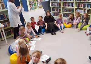Dzieci siedzą w kole i słuchają Pani Bibliotekarki, która tłumaczy jak należy wykonać pracę plastyczną