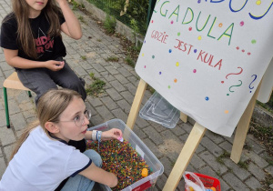 Dwie dziewczynki, jedna siedzi na krzesełku, druga losuje zagadkę z pojemnika z kulkami.
