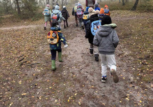 Dzieci podczas wycieczki biegną w lesie za przewodniczką.