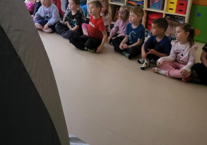 Grupa dzieci siedzi w siadzie skrzyżnym przed kinem sferycznym.