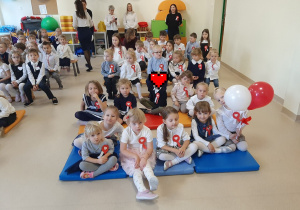 Dzieci ubrane w stroje galowe siedzą na materacach i ławkach