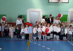 Dzieci ubrane w stroje galowe ustawiają się w dwuszeregu na sali gimnastycznej i czekają na odśpiewanie Hymnu