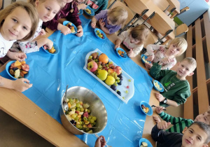 Dzieci z grupy Niebieskiej pozują przy wspólnym stole. Każde dziecko ma przed sobą miseczkę z sałatką owocową.