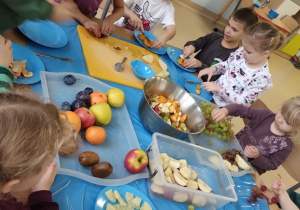 Dzieci z grupy Niebieskiej siedząc przy wspólnym stole kroją owoce na sałatkę owocową.
