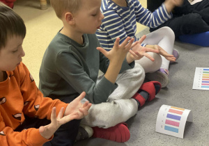 Dzieci siedzą na dywanie z rozłożoną klawiaturą dźwięków gamy C-durna i liczą wygrywane dźwięki na palcach.