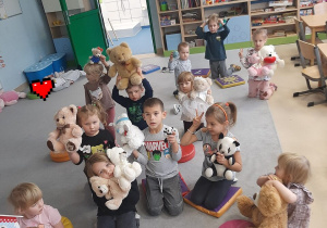 Dzieci siedzą na dywanie i unoszą pluszaki do góry