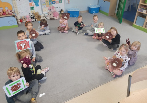 Dzieci siedzą w kole i unoszą do góry prace plastyczne Misie