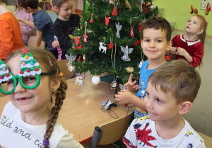 Dzieci ubierają choinkę, dziewczynka pozuje w świątecznych okularach