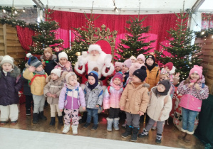 Dzieci w grupie pozują z Mikołajem na tle choinek
