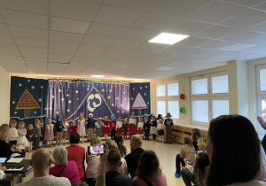 Dzieci z grupy Niebieskiej stoją w sali gimnastycznej na tle świątecznej dekoracji