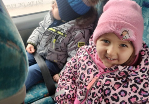 Dziewczynka i chłopiec siedzą w autokarze, chłopiec patrzy przez okno, dziewczynka uśmiecha się
