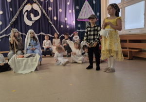 Na pierwszym planie chłopiec i dziewczynka jako Jaś i Małgosia, nieco dalej na krzesłach siedzą dzieci przebrane za Maryję i Józefa, za nimi grupa dzieci siedzi na ławce.