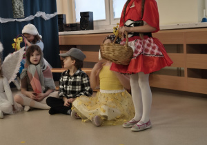 Dziewczynka przebrana za Czerwonego Kapturka, z boku siedzi trójka dzieci.