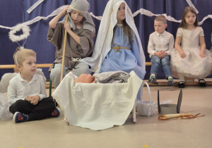 Na pierwszym planie siedzi chłopiec przebrany za Józefa i dziewczynka w stroju Maryi. Z lewej strony siedzi chłopiec, na drugim planie po lewej stronie siedzi dwoje dzieci.