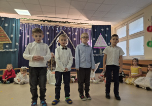 Na pierwszym planie stoi czterech chłopców, z tyłu siedzą dzieci.