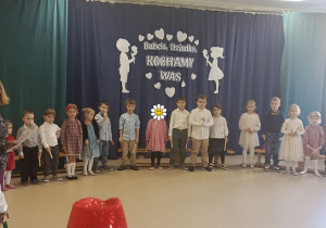 Dzieci stoją na tle dekoracji z okazji Dnia Babci i Dziadka i śpiewają okolicznościowe piosenki.