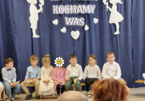 Dzieci siedzą na ławeczce na tle dekoracji z okazji Dnia Babci i Dziadka.