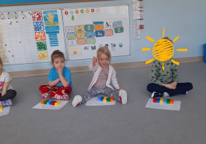 Dzieci siedzą na dywanie w kole, dziewczynka rozdaje dzieciom zestawy klocków LEGO Duplo w 6 kolorach