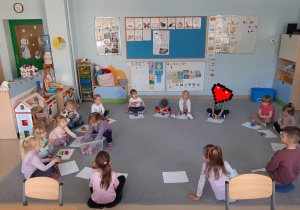 Dzieci siedzą na dywanie w kole, dziewczynka rozdaje dzieciom zestawy klocków LEGO Duplo w 6 kolorach