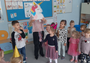 Dzieci ustawione w półkolu, Pani trzyma Piniatę Jednorożca w górze, dziewczynka pociąga za wstążeczki