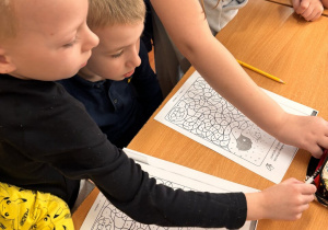 Dwóch chłopców, przedszkolaków podczas lekcji siedzi w ławce i koloruje obrazek z pomocą ucznia.