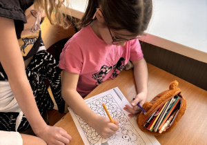 Dziewczynka siedzi w szkolnej ławce i koloruje obrazek z pomocą starszej uczennicy