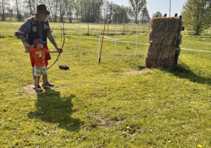 Chłopiec z pomocą Indianina uczy się strzelać z łuku w kierunku słomianego słupa.