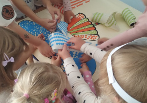 Dzieci w udekorowanych przez siebie opaskach na głowie współpracują podczas naklejania kropek na karton